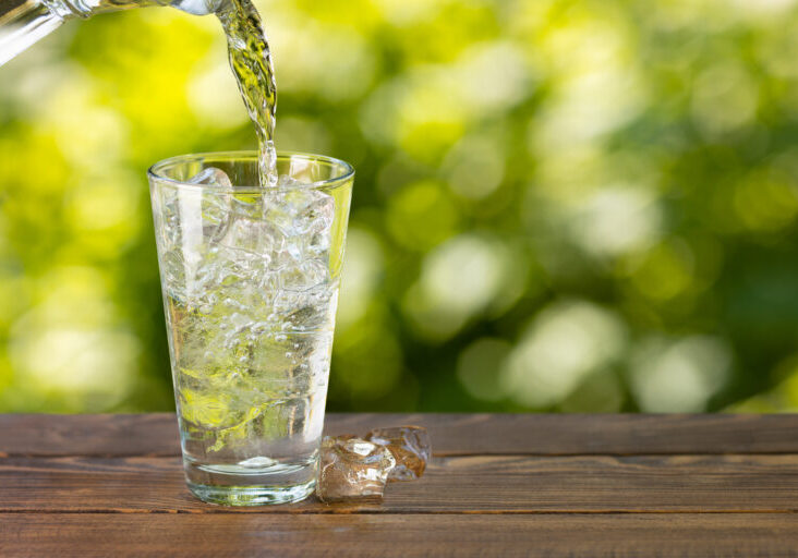 water from jug pouring into glass with ice cubes on wooden table outdoors